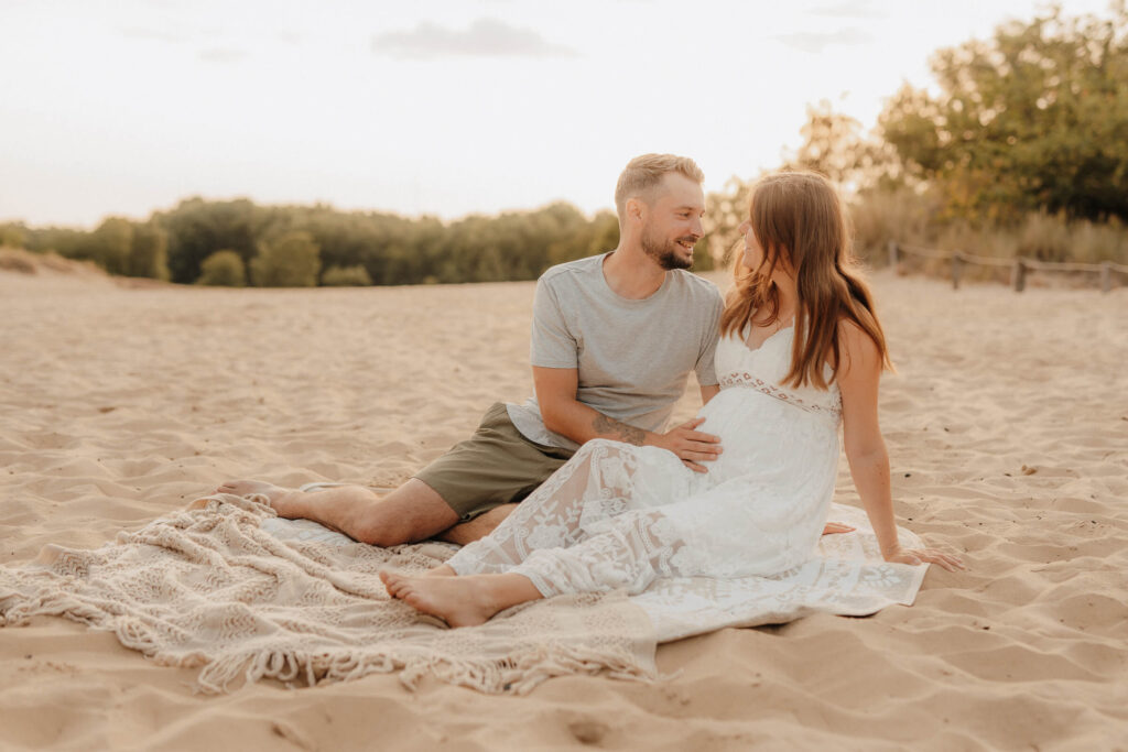 babybauchshooting boberger duenen, schwangere und partner sitzen auf decke und laecheln sich an
