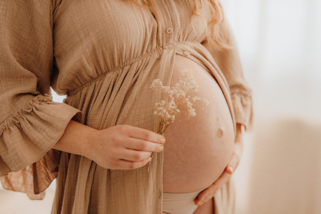 babybauchshooting zuhause, schwangere beruehrt ihren bauch und haelt trockenblumen in der hand