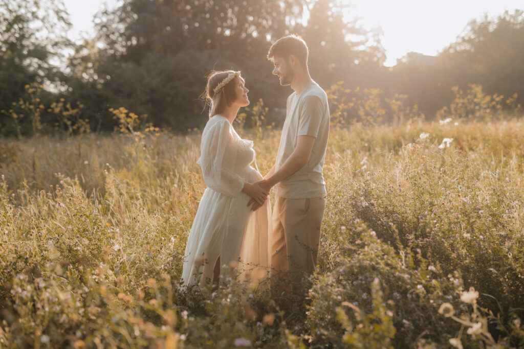 babybauchshooting auf wilder wiese, schwangere und partner stehen sich gegenueber und beruehren den babybauch
