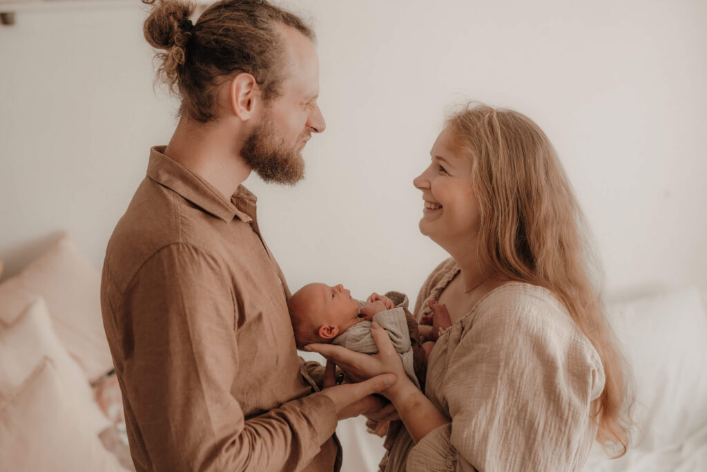 newbornshooting, baby liegt in mamas und papas armen, mama und papa gucken sich lustig an